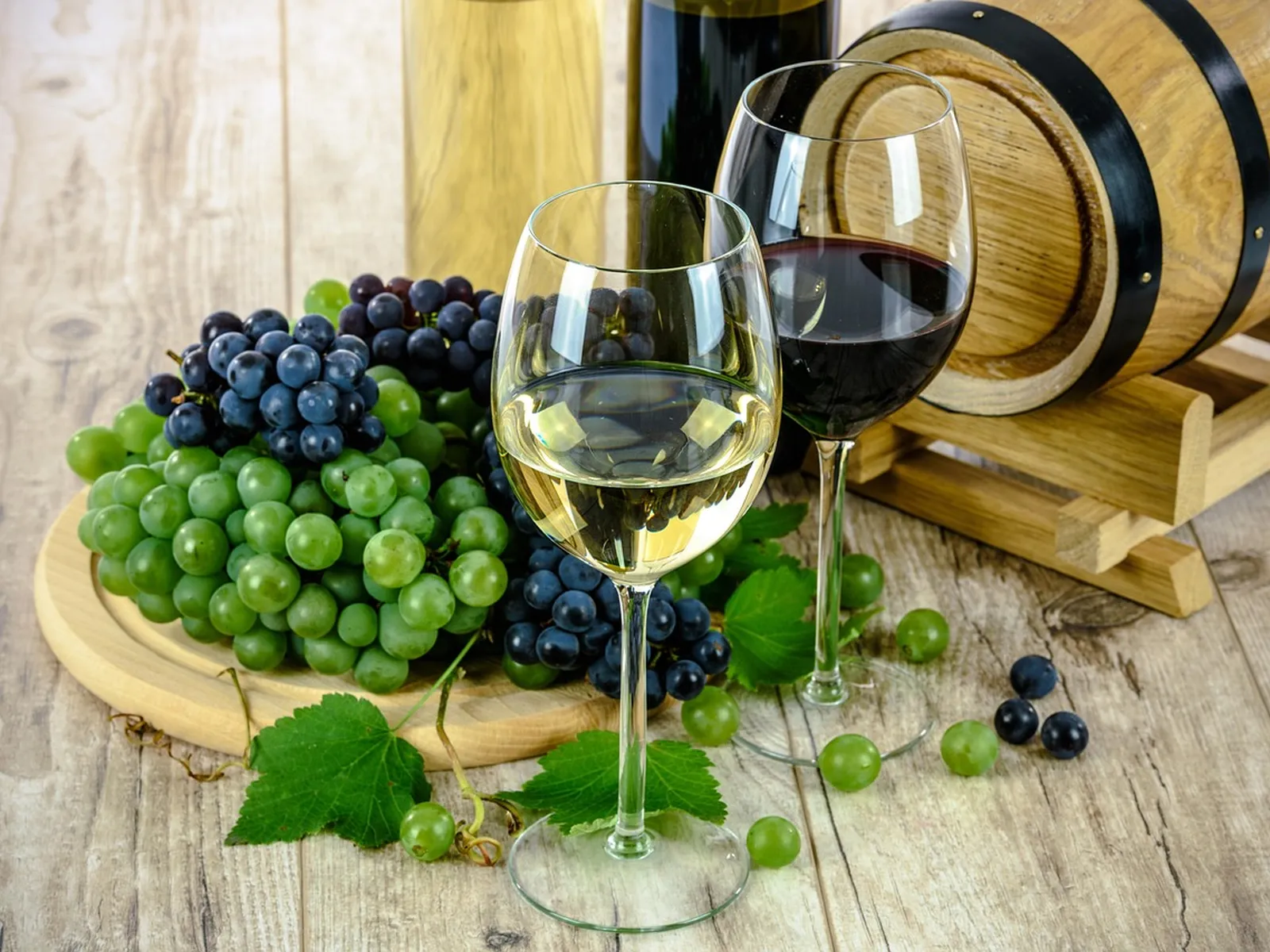 A glass of white and a glass of red wine beside bunches of grapes and a barrel